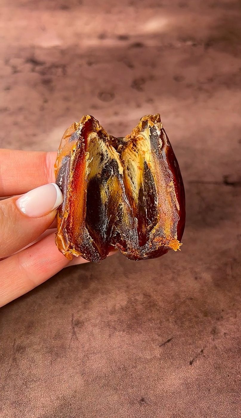 A hand with a manicured nail holds a halved date, revealing its sticky, fibrous interior. The background is a soft, brown surface.