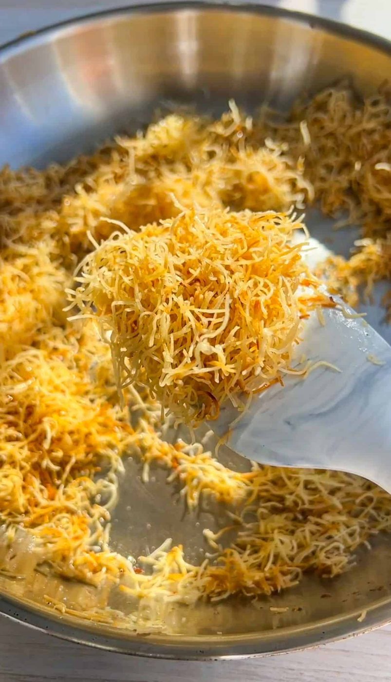 A close-up of a pan with golden, toasted shredded phyllo dough. A white spatula is lifting some of the crispy strands, showcasing their texture and color. The background is softly blurred, focusing on the phyllo dough.