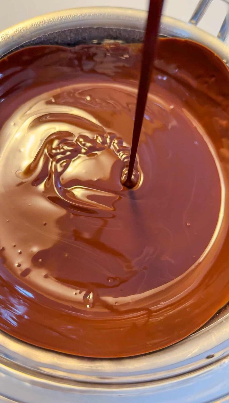 A stream of liquid chocolate pours into a bowl filled with melted chocolate, creating swirling patterns on the glossy surface.
