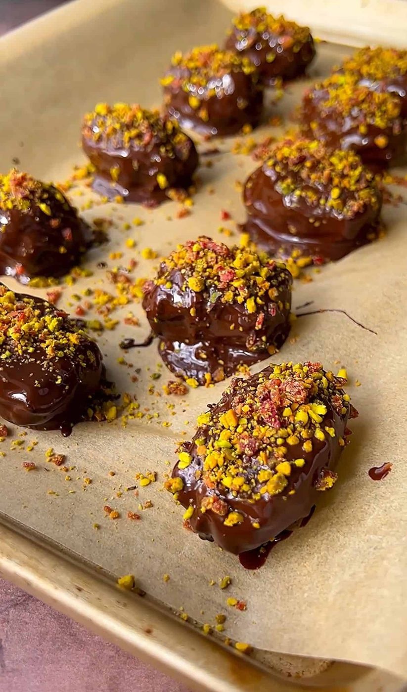 Dubai chocolate dates topped with crushed nuts and colorful sprinkles are arranged on a parchment-lined baking sheet. The treats are glossy from the chocolate coating and appear freshly made.