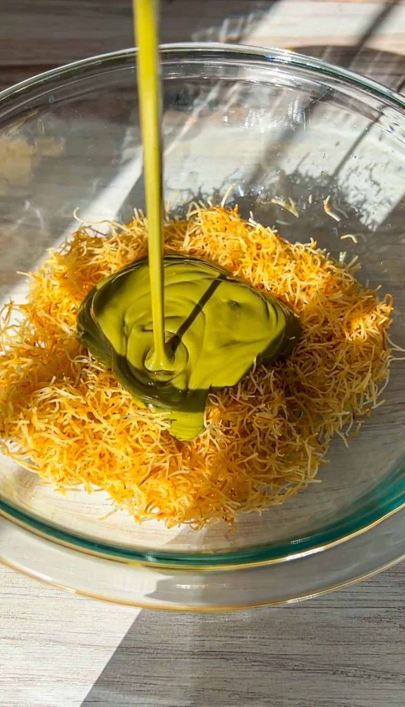 Shredded pastry dough is placed in a glass bowl with a green paste on top. A stream of golden liquid is being poured over the mixture. Bright sunlight is casting shadows on the surface.