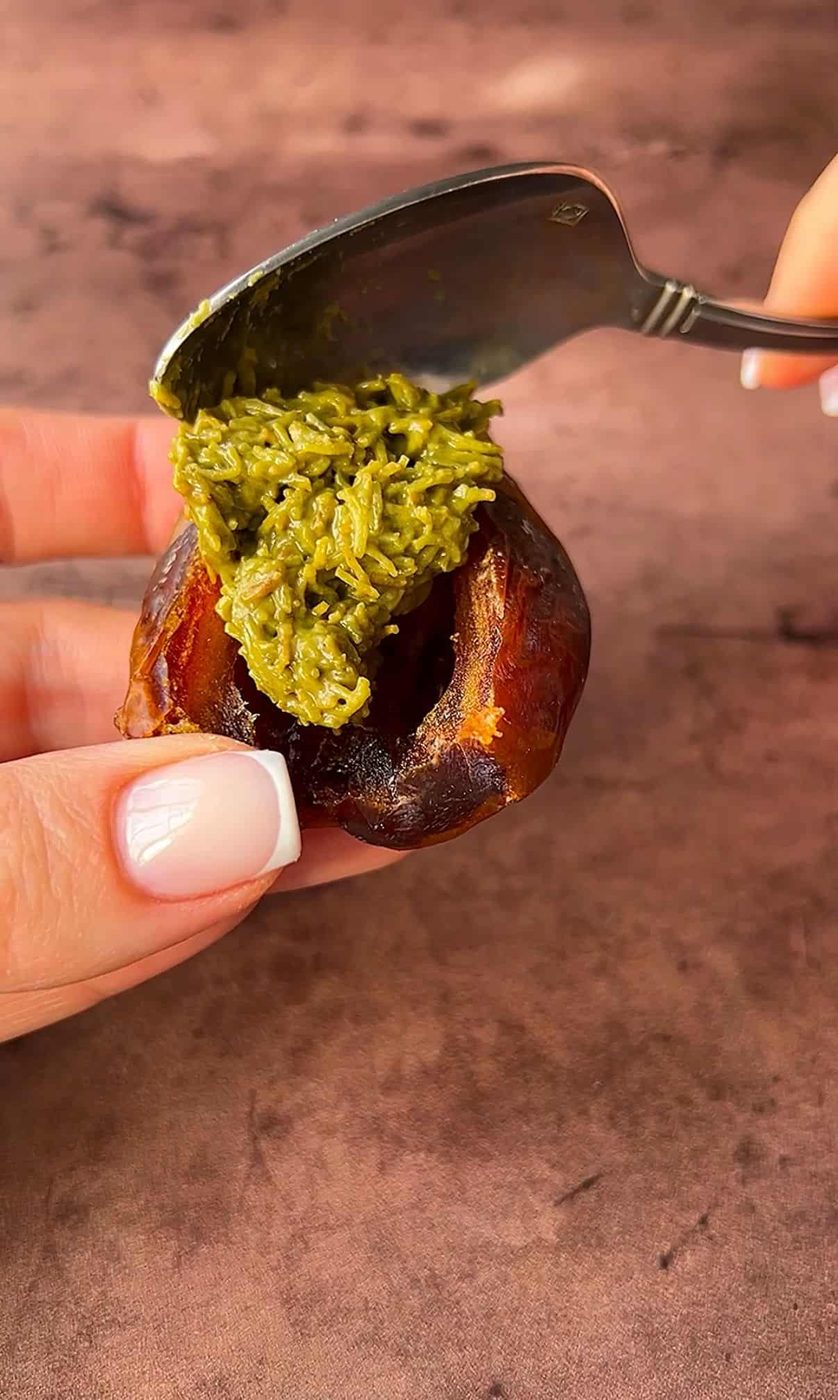 A hand is holding a date, and another hand is using a spoon to fill it with a green paste. The background is a brown surface.