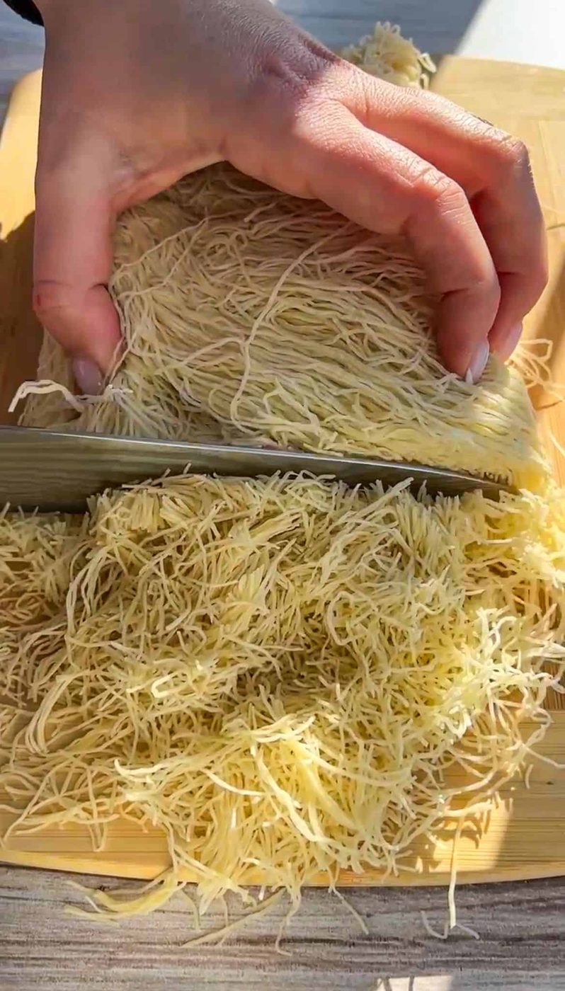 A hand holding a knife slices through a block of fine shredded dough, possibly kataifi, on a wooden cutting board placed on a light-colored table. The dough is spread out as it's being cut.