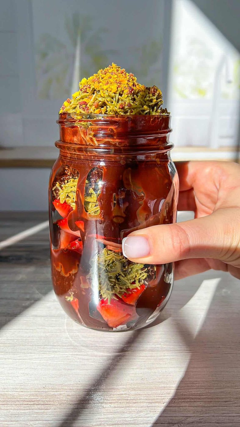 A hand holds a jar filled with layered Dubai Strawberry Cup, including strawberries and chocolate sauce, topped with green pistachio crumbs. Sunlight casts shadows on the textured wooden surface below.