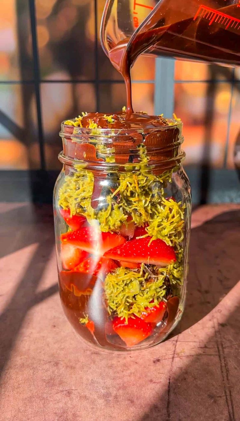 A jar filled with sliced strawberries and green shredded coconut is being topped with a pour of melted chocolate from a measuring cup. The mixture appears rich and colorful, with sunlight streaming in from a window behind.