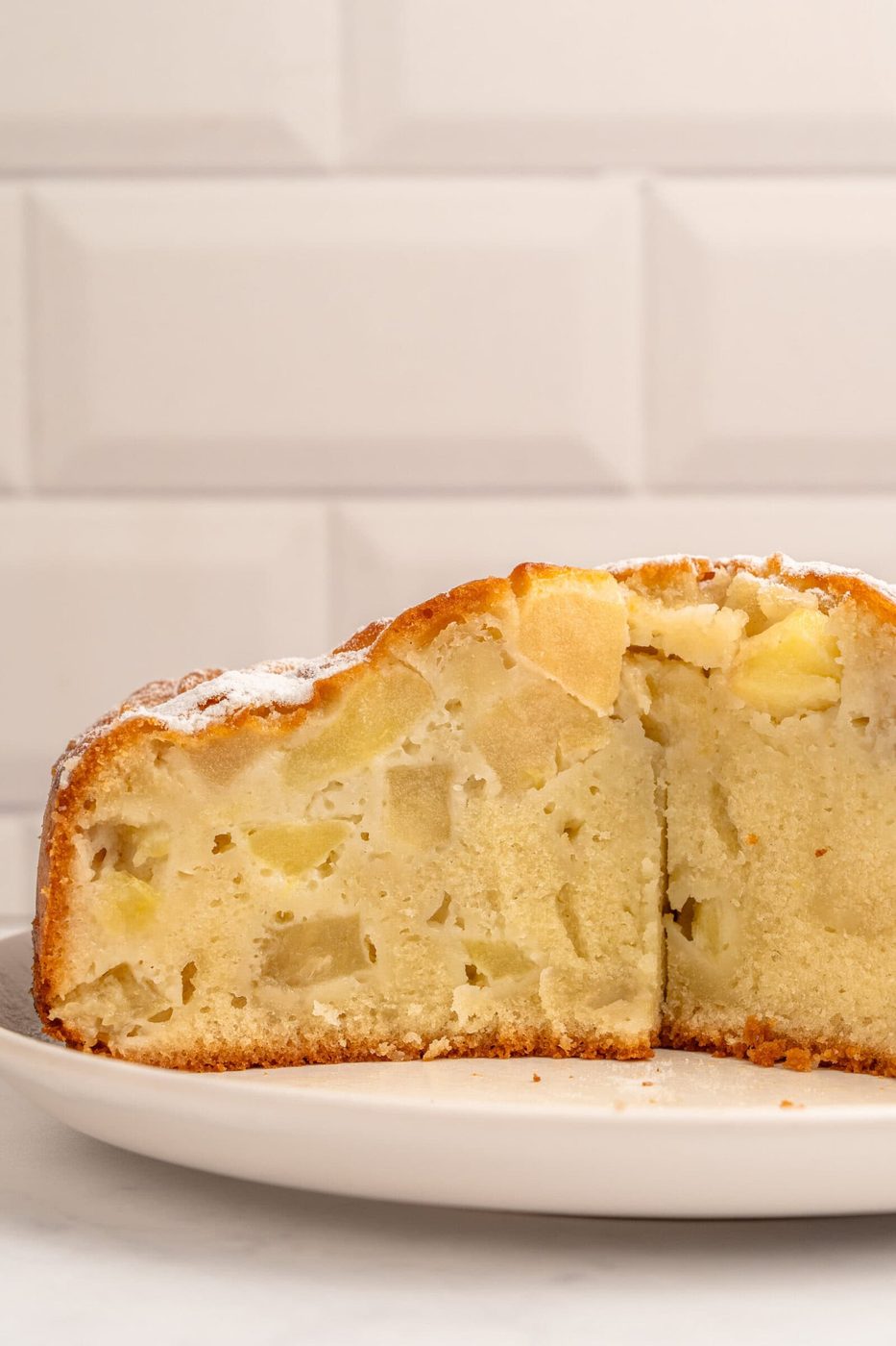 A close-up of a sliced apple cake on a white plate, showing its moist, dense texture and chunks of apple, with a light dusting of powdered sugar on top. A white tiled background is visible.