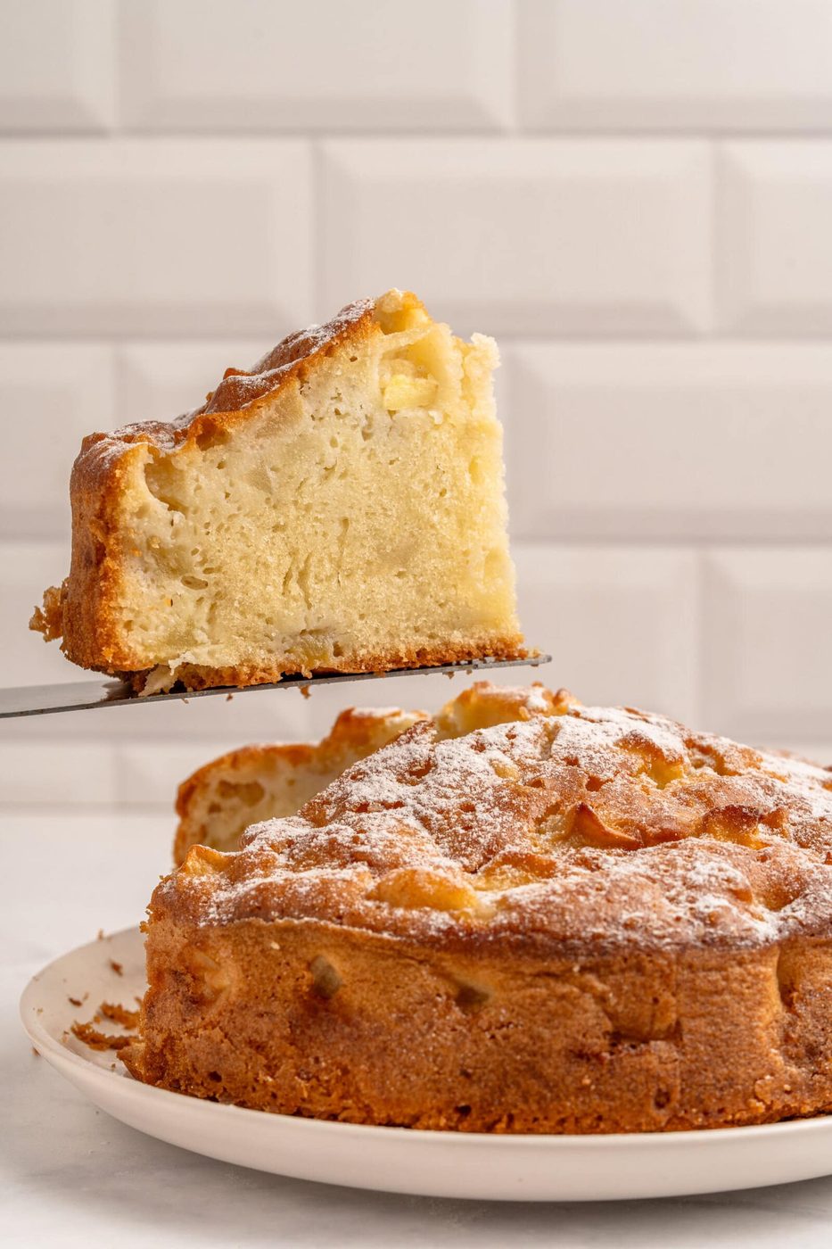 A slice of homemade cake is being lifted from a whole cake on a white plate. The cake has a golden brown crust and is dusted with powdered sugar. The background features a white tiled wall.