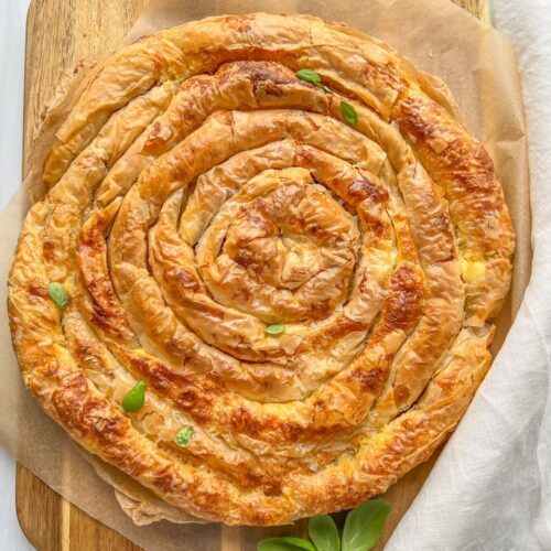 A large, golden-brown Banitsa spiral pie with flaky phyllo pastry sits on parchment paper atop a wooden board, garnished with a few fresh green basil leaves.