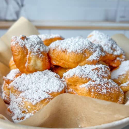 A bowl lined with parchment paper filled with golden, fluffy beignets and Mekitsi, generously dusted with powdered sugar, set on a white surface with a blurred kitchen background.