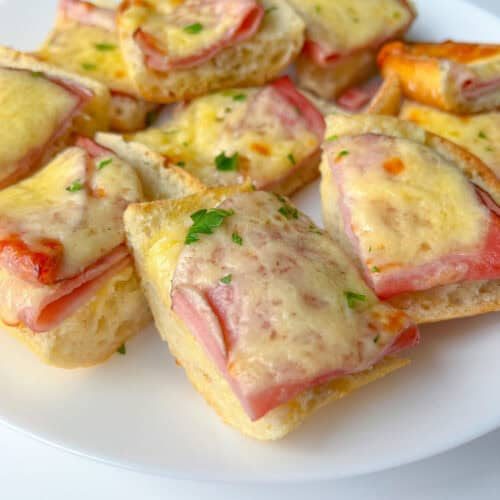 Slices of bread topped with ham and melted cheese create classic Ham and Cheese Sandwiches, garnished with chopped parsley and beautifully arranged on a white plate.