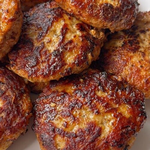A close-up of several golden-brown, pan-fried sausage patties stacked on a white plate. The crispy, caramelized exterior and juicy texture make them perfect for anyone looking for an Easy Meatball Recipe alternative.