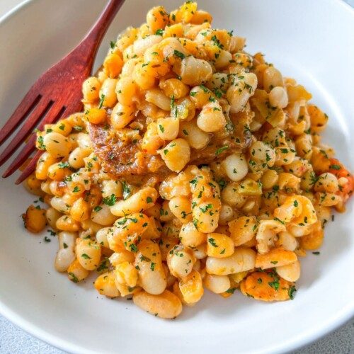A white bowl filled with seasoned white beans garnished with chopped herbs, served with a reddish-brown fork on the side.
