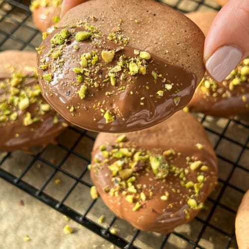 A hand holding a round cookie dipped in chocolate and sprinkled with crushed pistachios, with similar cookies resting on a wire cooling rack in the background.