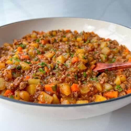 A close-up of a bowl filled with a chunky stew resembling Moussaka with Potatoes, featuring ground meat, diced potatoes, carrots, and green herbs, with a wooden spoon resting inside. The dish appears hearty and freshly prepared.