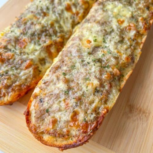 Two pieces of garlic bread topped with melted cheese and herbs, reminiscent of Bulgarian Meat Sandwiches (Prinzesi), rest on a wooden cutting board. The bread appears golden and crisp around the edges.