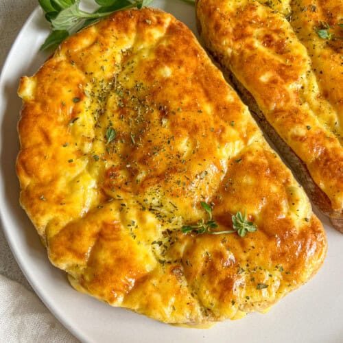 Two pieces of golden, freshly baked flatbread garnished with herbs are served on a white plate, accompanied by a sprig of fresh green oregano, reminiscent of flavors found in Feta Egg Toast.