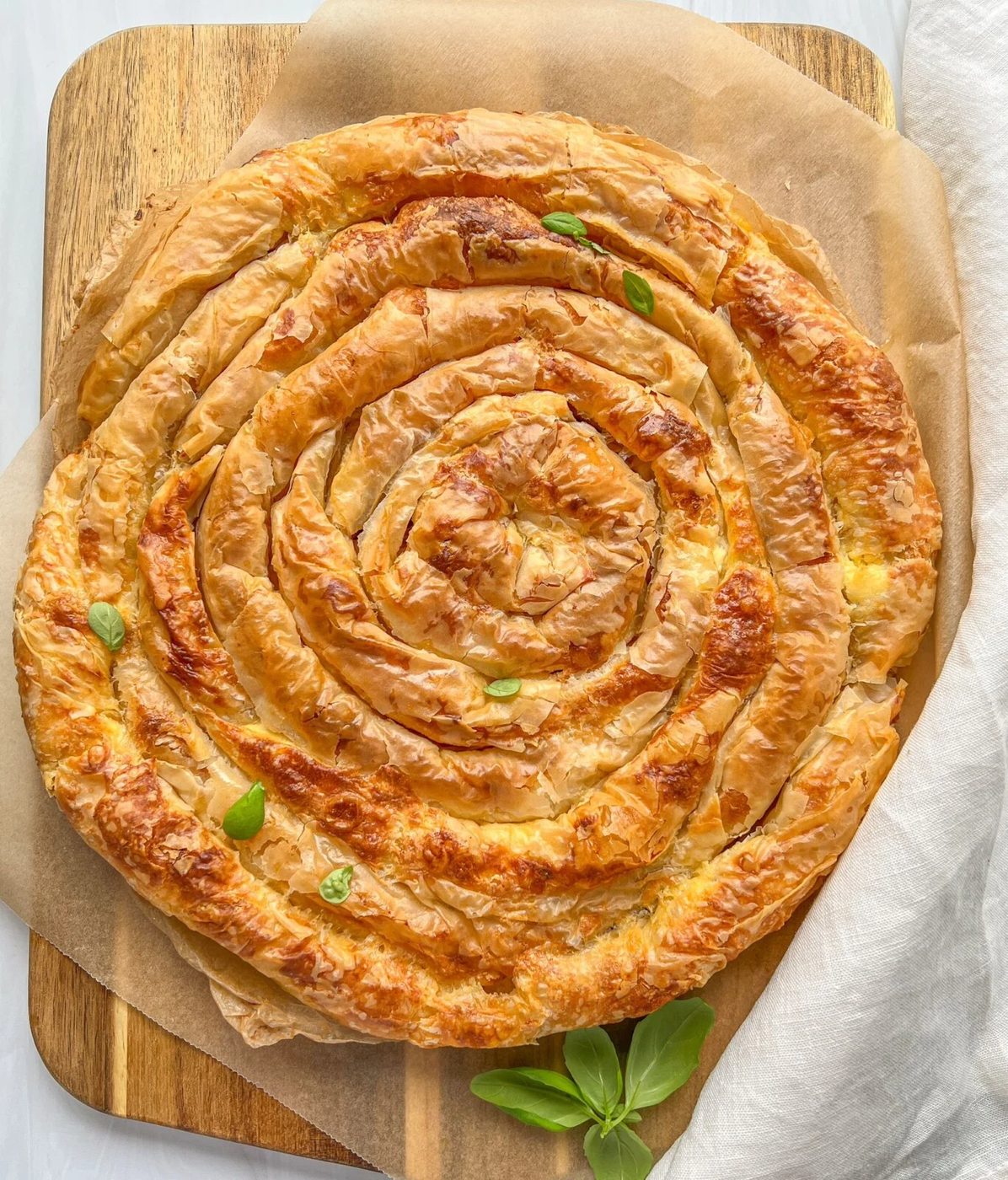 A golden-brown easy Bulgarian banitsa on a wooden cutting board, lined with parchment paper. The pastry is flaky and crisp, with a few basil leaves scattered on top for garnish. A white cloth is partially visible on the side.