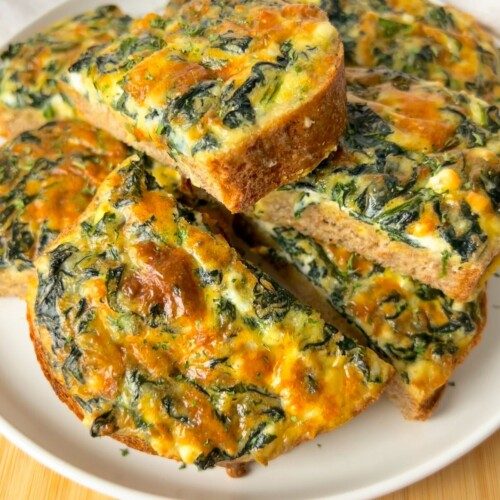 Slices of baked spinach and cheese frittata are arranged on a white plate, showing a golden-brown crust and a colorful mixture of spinach and melted cheese inside.