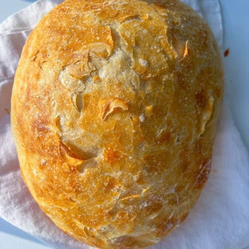 A round, golden-brown loaf of No Knead Bread rests on a folded white cloth, boasting a crisp, textured surface and rustic appearance.