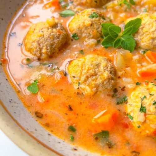 A close-up of a bowl of soup with meatballs, diced carrots, rice, herbs, and a creamy orange broth, garnished with fresh green herbs.