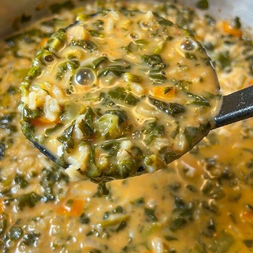 A close-up of a creamy spinach soup recipe with chopped greens, carrots, and rice, being scooped from a stainless steel pot with a black ladle.