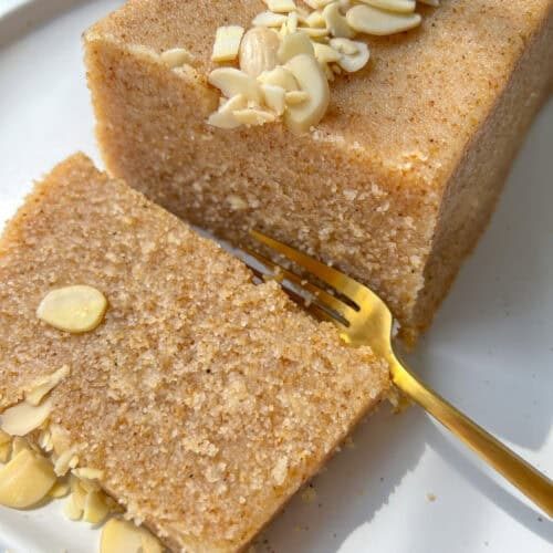 A close-up of a rectangular slice of Semolina Cake topped with sliced almonds on a white plate. A gold fork rests beside the cake, with one piece of the Semolina Cake partially cut and ready to eat.