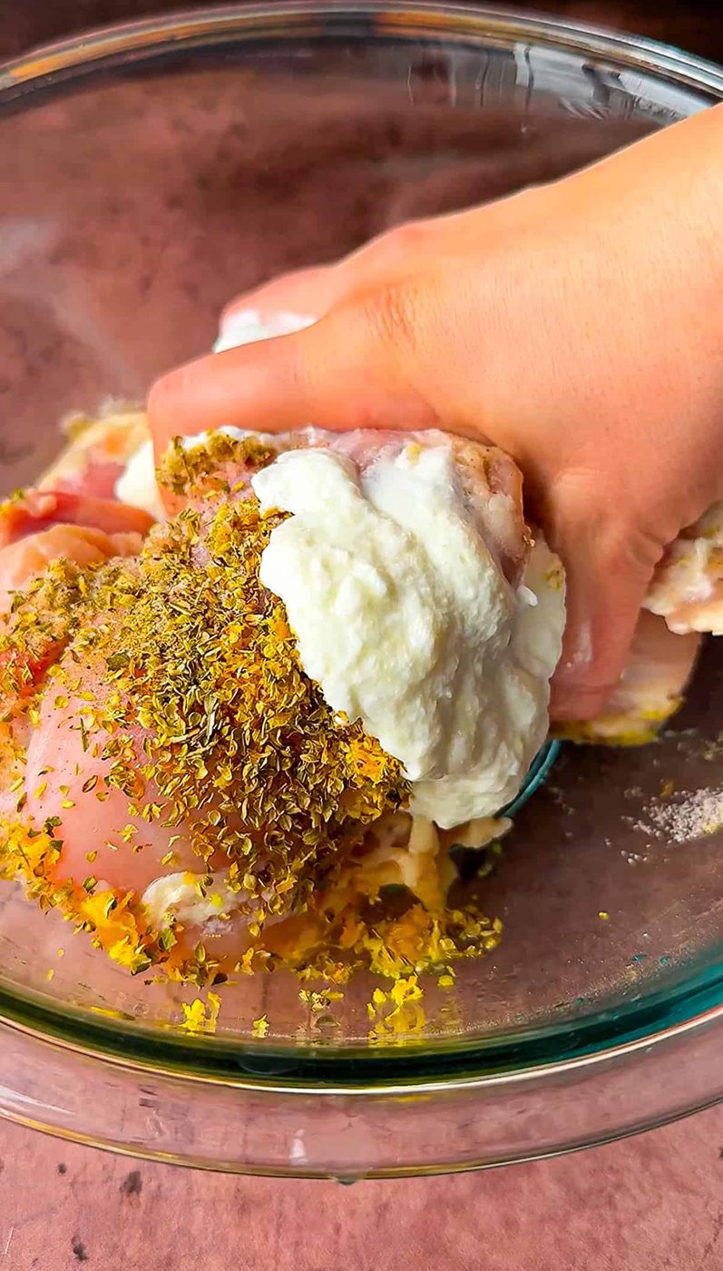 Hand mixing raw chicken pieces with a marinade of yogurt, herbs, and spices in a glass bowl.