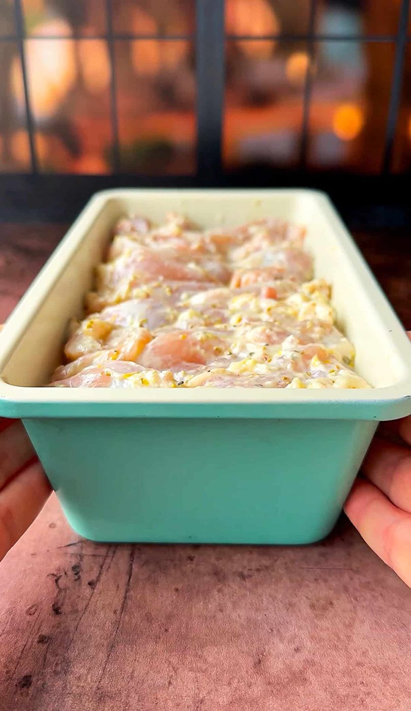 Hands holding a light blue loaf pan filled with marinated thighs, ready to be cooked. The background shows a warm, softly lit kitchen.