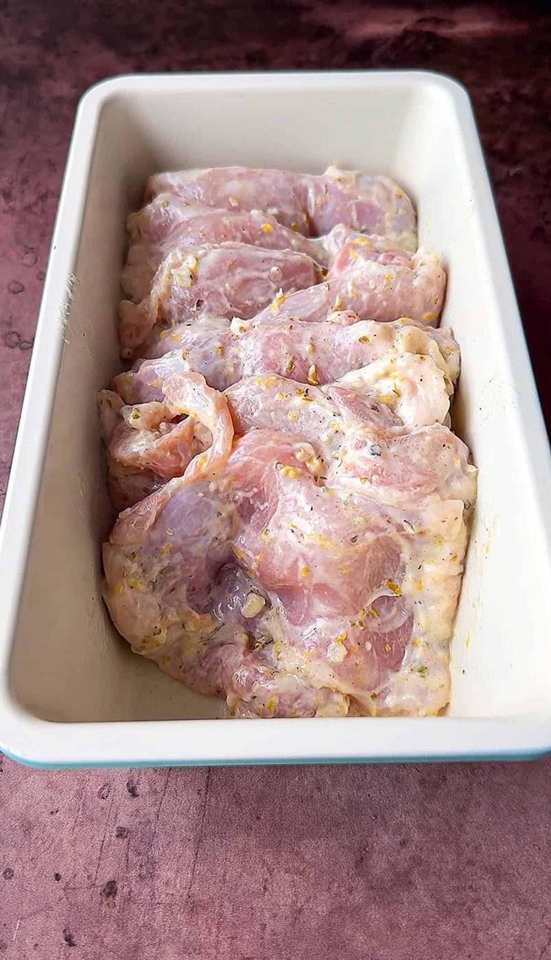 Raw chicken marinating in a rectangular pan, covered with a mixture of herbs and spices. The marinade appears to contain visible pieces of garlic and other seasonings. The pan is placed on a dark, textured surface.