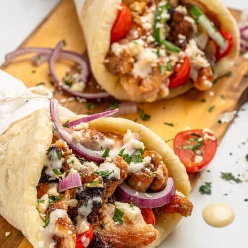 Two gyros filled with grilled meat, fresh tomatoes, red onion slices, crumbled feta, and herbs are served on a wooden board. A creamy sauce is drizzled on top, and the gyros are wrapped in pita bread.