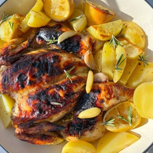 Spatchcock Greek Lemon Chicken with crispy, browned skin is served alongside golden potato wedges, lemon halves, garlic cloves, and sprigs of rosemary in a white dish.