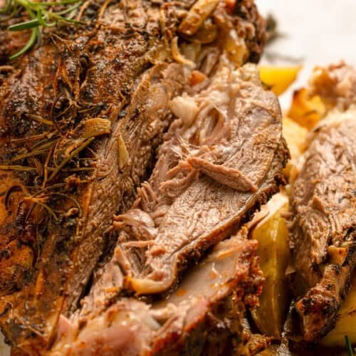 Close-up of a juicy, roasted lamb shoulder garnished with herbs. The meat is tender and shredded, with crispy, seasoned edges and visible sprigs of rosemary.