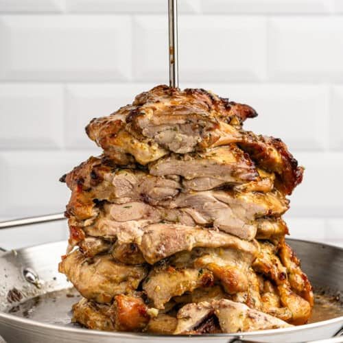 Stacked layers of marinated, grilled meat on a vertical skewer in a metal pan, set against a white tiled background.
