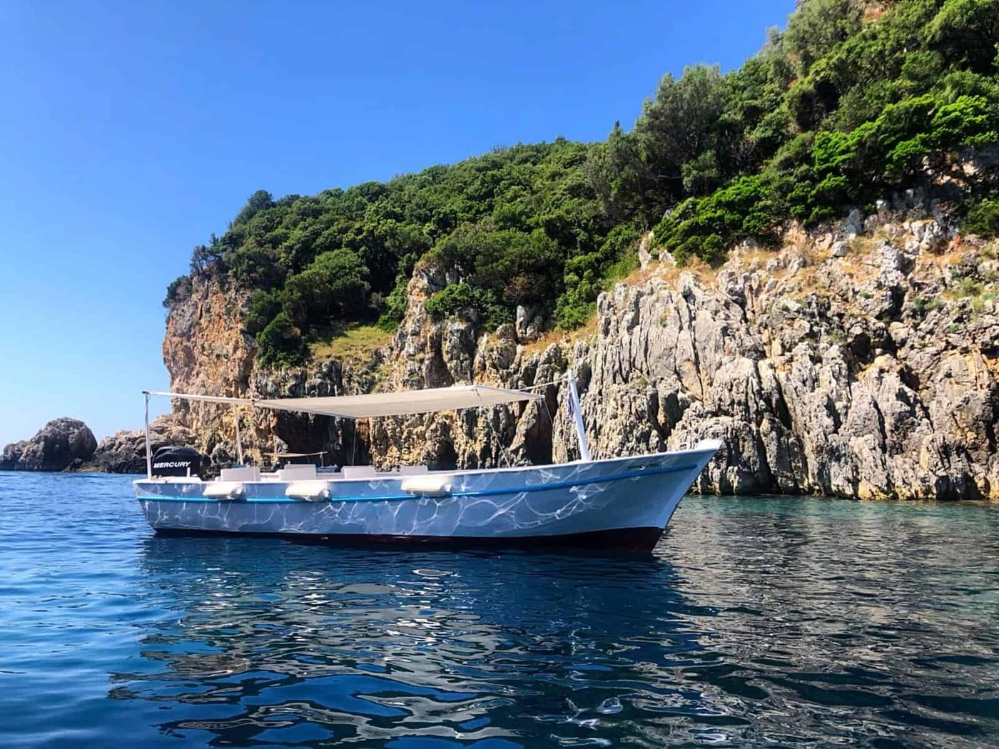 A boat with a beige canopy floats on clear blue water near a rocky cliff covered with green vegetation. The sky is clear and blue, creating a serene and picturesque coastal scene. This is the place where I learned Greek Recipes.