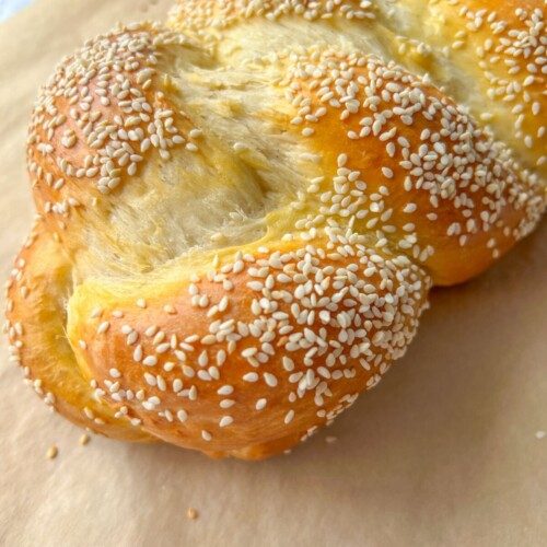 A close-up of a golden-brown braided Sweet Easter Bread, topped with sesame seeds and resting on a sheet of parchment paper.