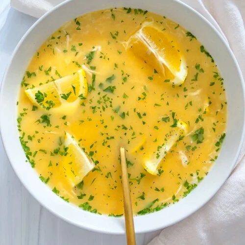 A bowl of yellow soup inspired by modern the cook recipes is garnished with parsley and lemon wedges, with a gold spoon resting inside. The bowl sits on a white surface beside a cream-colored cloth.