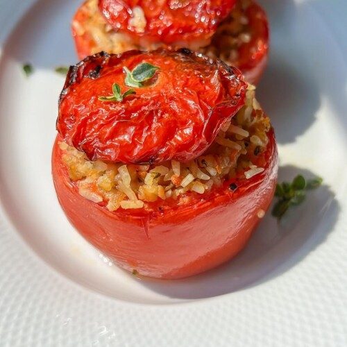 Three Stuffed Tomatoes baked with rice and herbs are arranged in a row on a white plate. Their grilled tops and fresh herb garnish catch the sunlight, creating an inviting scene.