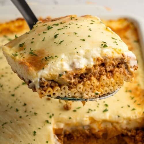 A close-up of a slice of pasta topped with melted cheese and garnish, being lifted from a baking dish. The pasta is layered with a meat sauce and creamy topping, with parsley sprinkled on top. The dish is set against a neutral background.
