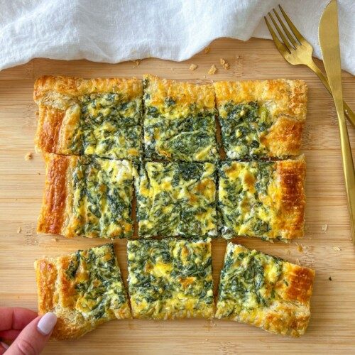 A hand holds a slice of savory Puff Pastry Strudel topped with a spinach and cheese mixture, cut into squares and arranged on a wooden board next to a gold fork and knife.