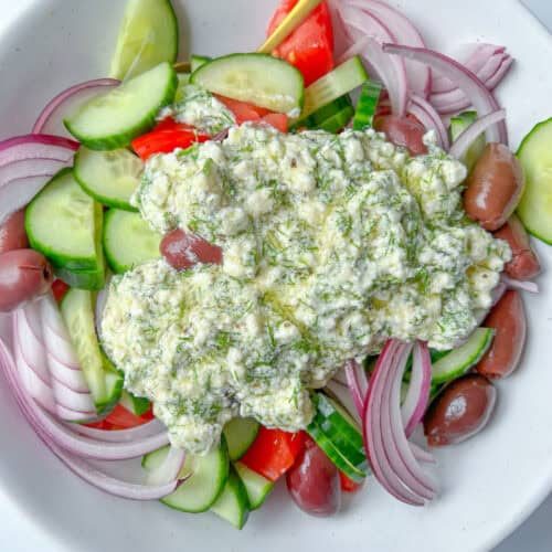 A bowl of Greek Feta Salad with sliced cucumbers, tomatoes, red onions, Kalamata olives, and a generous dollop of tzatziki sauce on top, with a spoon resting in the salad.