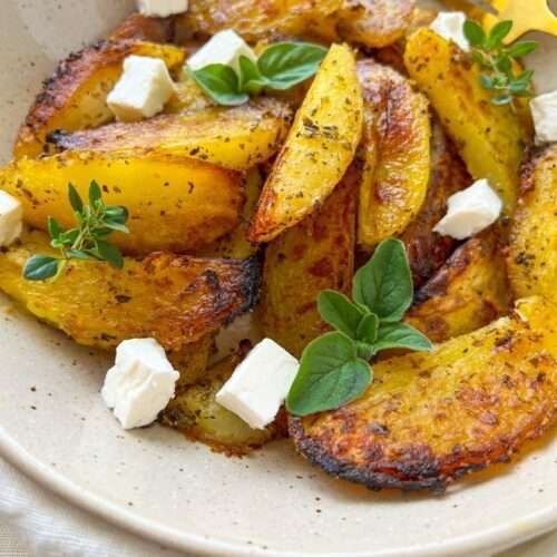A plate of golden, seasoned Greek Lemon Potatoes wedges garnished with fresh herbs and small cubes of white cheese, with a gold fork on the side.