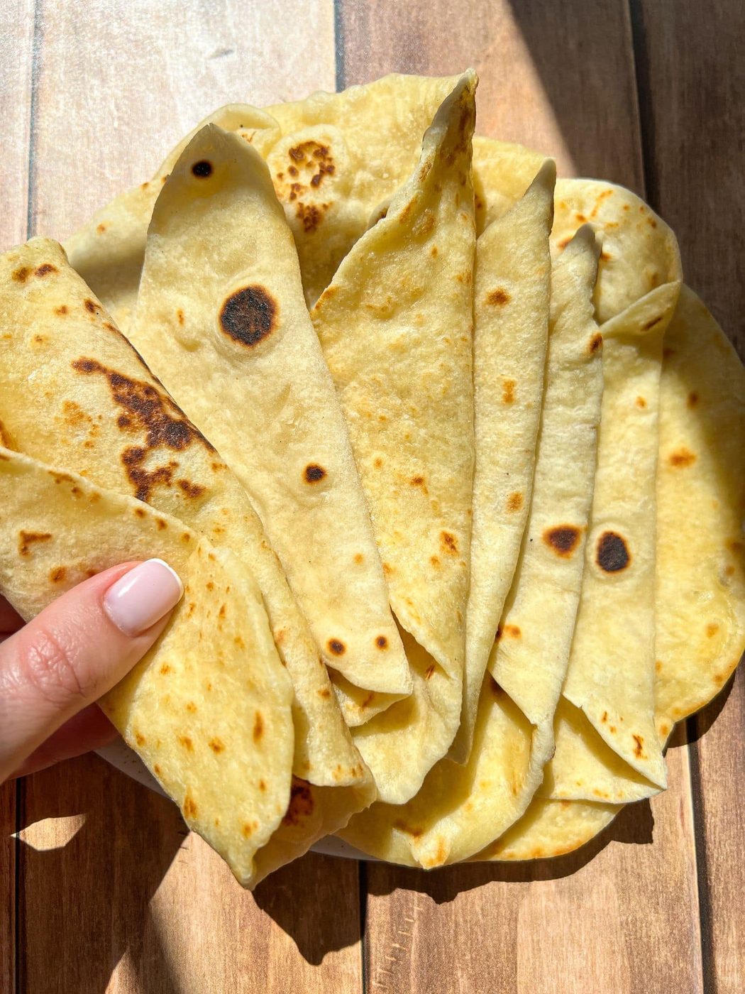 A hand holds several folded, golden-brown tortillas, showcasing their soft texture and slight charring. The tortillas are placed on a wooden surface, highlighted by bright sunlight.