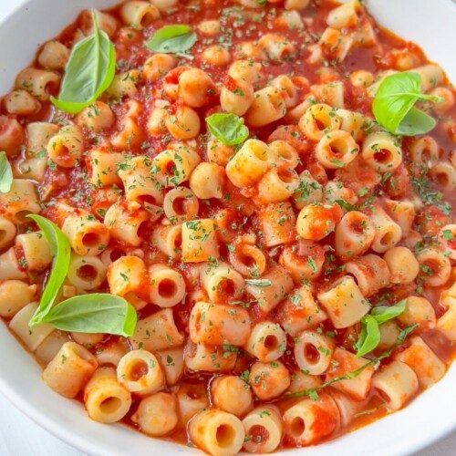 A white bowl filled with ditalini pasta in a rich tomato sauce, inspired by Pasta e Ceci, garnished with fresh basil leaves and sprinkled with chopped herbs.