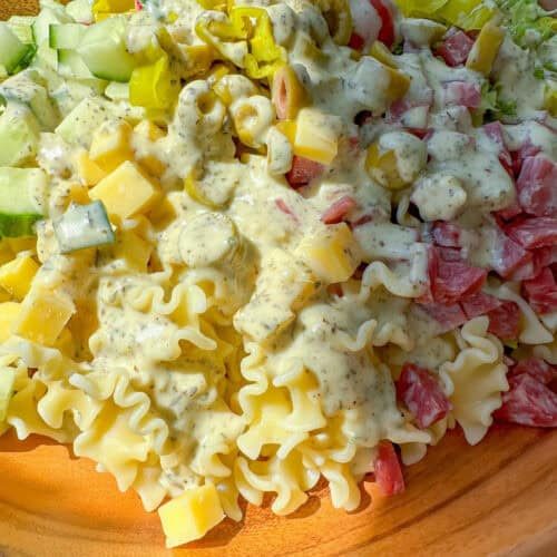A wooden bowl filled with Grinder Pasta Salad—pasta, diced tomatoes, cucumbers, cheese cubes, olives, pepperoncini, and lettuce—all topped with creamy Italian dressing.