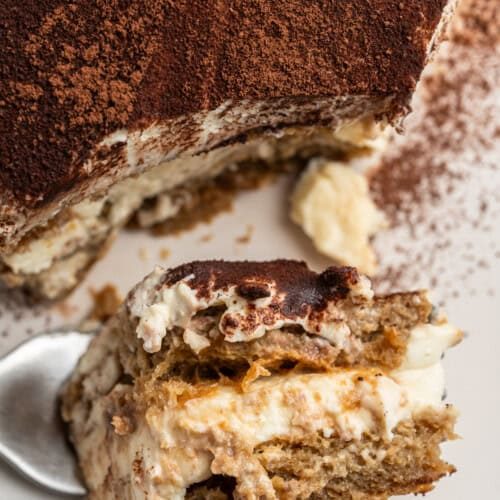 Close-up of a creamy tiramisu dessert on a white plate. A portion has been scooped out onto a spoon, revealing layers of espresso-soaked ladyfingers and mascarpone filling, dusted with cocoa powder.