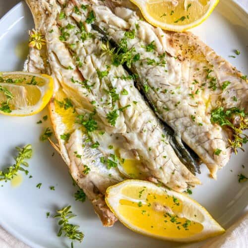 A plate with grilled fish garnished with chopped herbs and lemon wedges. The fish looks tender and seasoned, with olive oil drizzled on top. The lemons add a splash of color against the white plate.