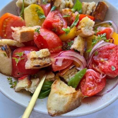 A bowl of Panzanella Salad with colorful tomato wedges, toasted bread chunks, sliced red onions, basil leaves, and a light dressing is served with two gold forks.