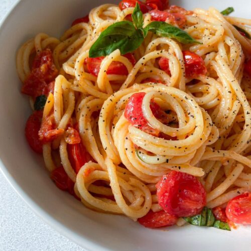 A bowl of Cherry Tomato Pasta with fresh basil and a sprinkle of black pepper, served in a white dish on a light surface.