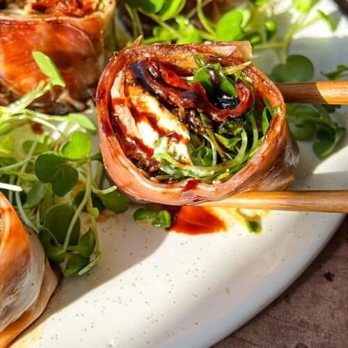 Close-up of Italian sushi recipe wrapped in prosciutto, filled with greens, and drizzled with a dark sauce on a white plate. They're accompanied by small leafy greens, with chopsticks on the side. The setting is warmly lit.