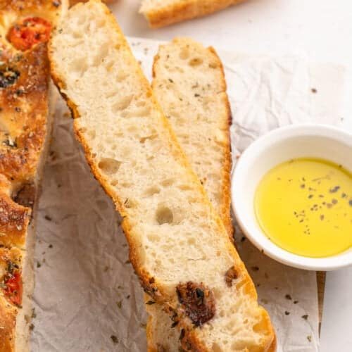 Slices of freshly baked focaccia bread with herbs and tomatoes are laid on parchment paper. A small bowl of olive oil is on the side for dipping.